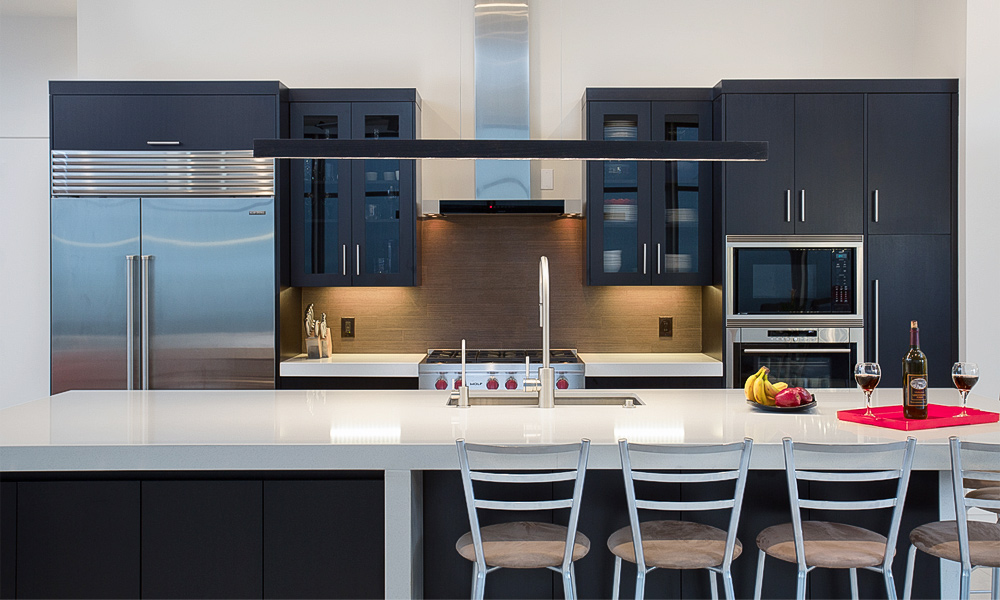 A close-up of a linear kitchen featuring dark lacquered base cabinets, with a white countertop on the central island. The sleek, glossy finish of the dark cabinets contrasts beautifully with the clean, bright white of the island's surface, emphasizing the modern, minimalist aesthetic of the space. The arrangement showcases the smooth lines and functional design elements of this contemporary kitchen layout.
